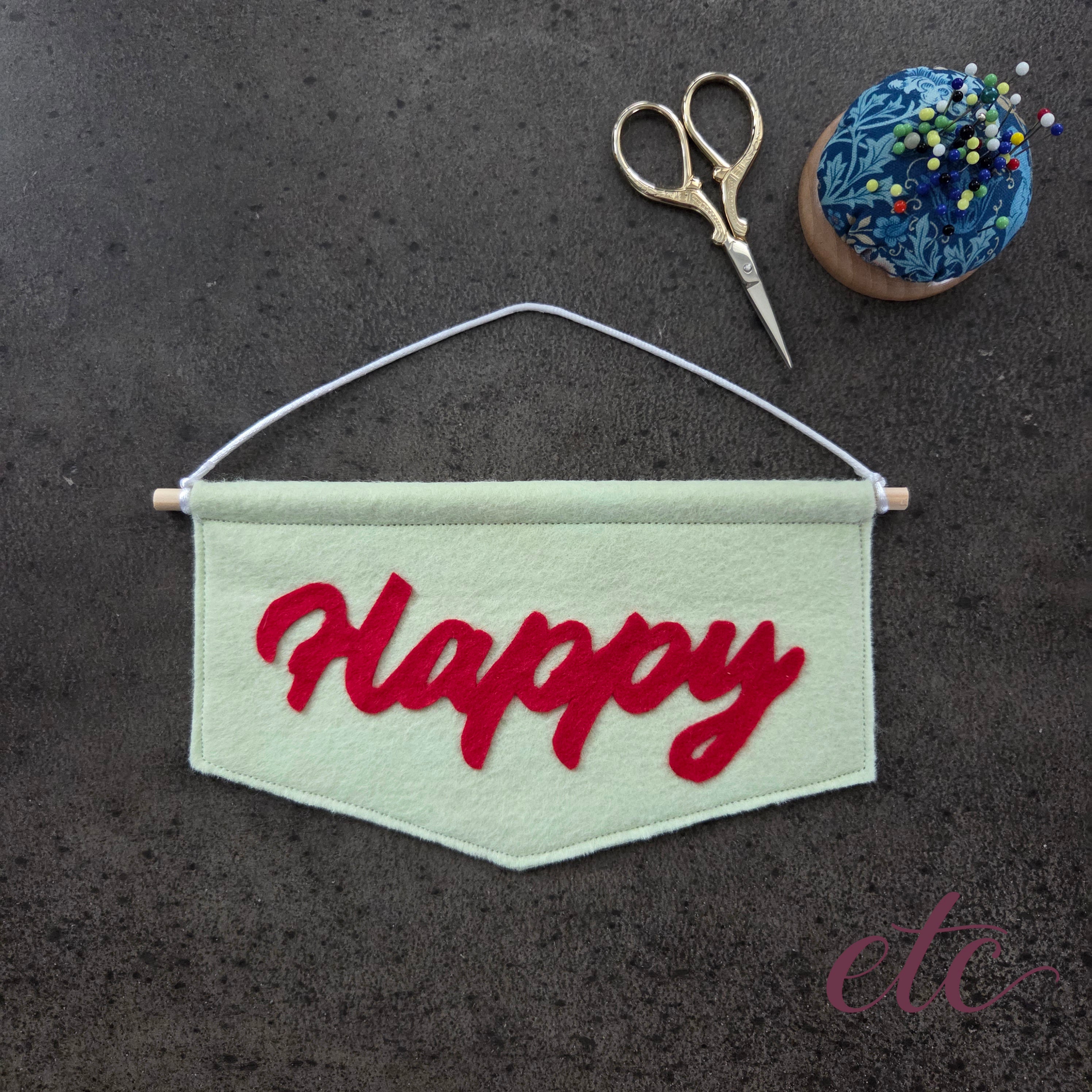 A handmade felt banner with the word 'Happy' in red cursive lettering, hanging from a wooden dowel with white string. The banner is styled on a dark textured surface with gold-handled scissors and a blue pincushion pin holder in the background.