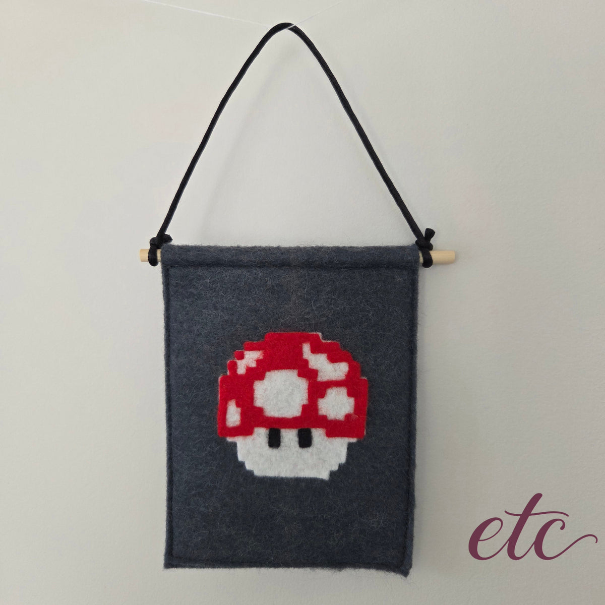 A dark gray felt wall hanging with a black hanging cord and wooden dowel, featuring a pixel-art Mario Bros red and white mushroom design in the center.