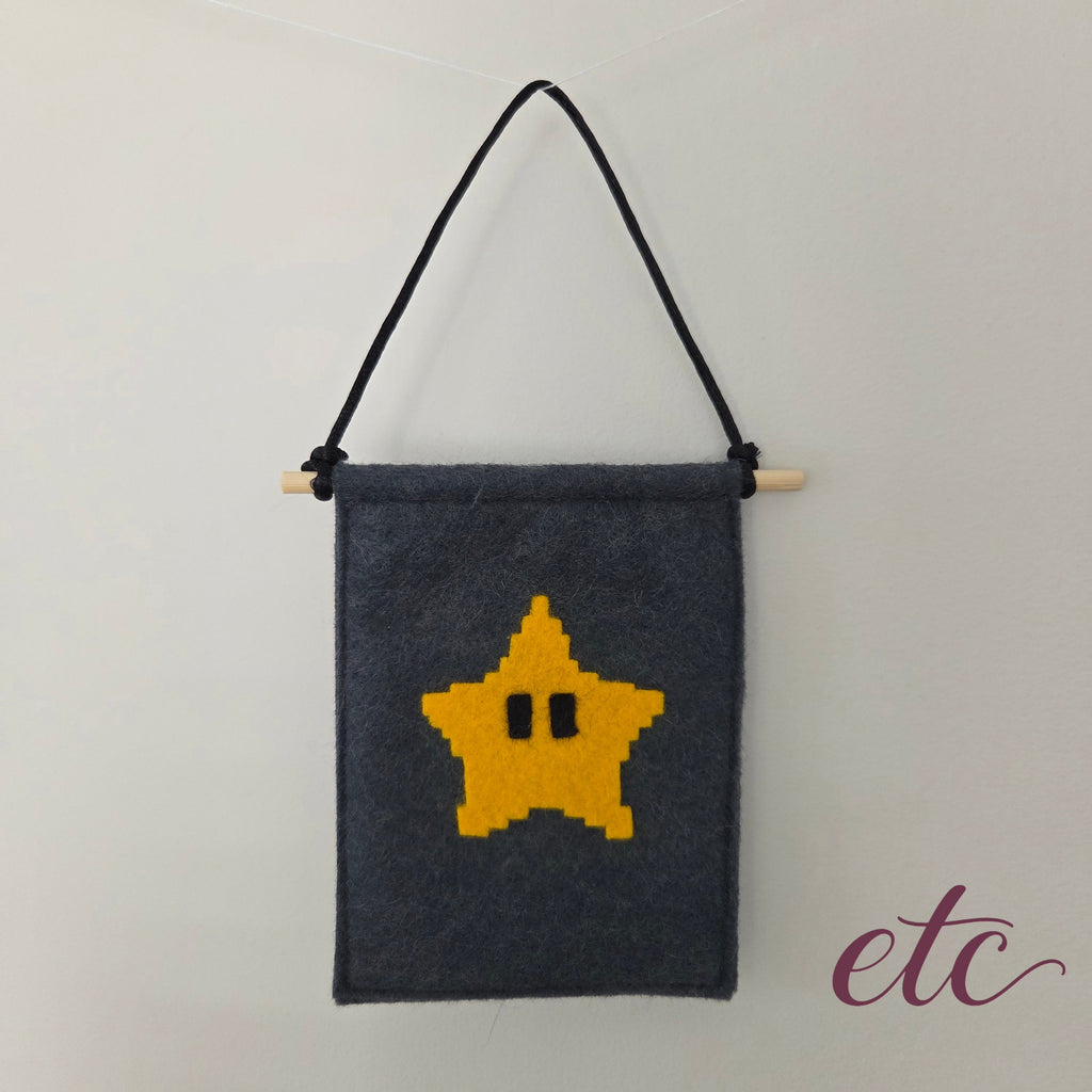 A dark gray felt wall hanging with a yellow pixelated star on the front, suspended from a light wooden dowel with a black hanging cord, shown against a plain light background.