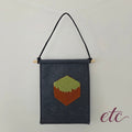 A dark gray felt wall hanging with a black hanging cord and a small pixel-art MineCraft block motif in green and brown, suspended against a plain light background.