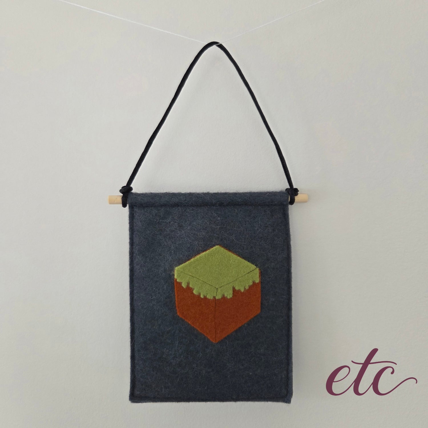 A dark gray felt wall hanging with a black hanging cord and a small pixel-art MineCraft block motif in green and brown, suspended against a plain light background.
