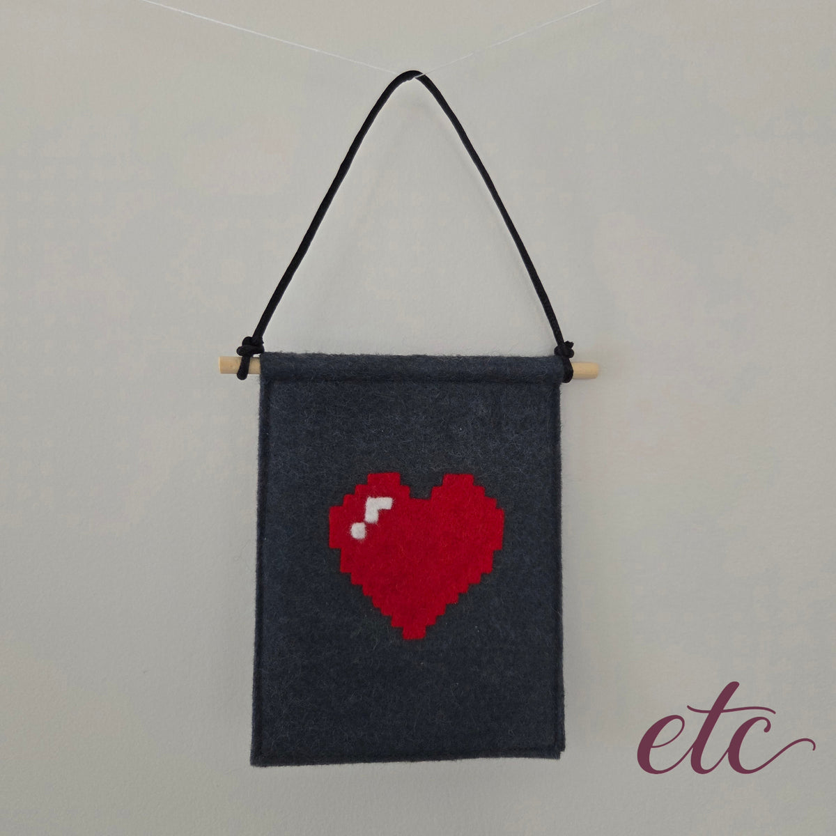 A dark gray felt wall hanging with a wooden dowel and black hanging cord, featuring a pixelated red heart with a small white detail on the front.