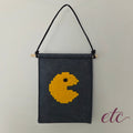 A dark gray felt wall hanging featuring a yellow pixelated Pac-Man design, suspended from a wooden dowel with a black hanging cord, against a plain light background.