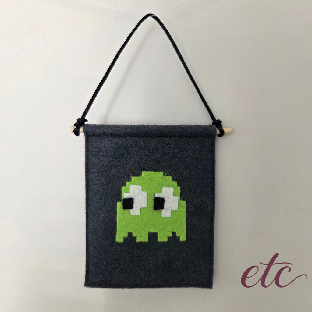 A dark charcoal gray felt wall hanging with a black hanging cord and a light wooden dowel, featuring a pixelated PacMan green ghost design on the front.