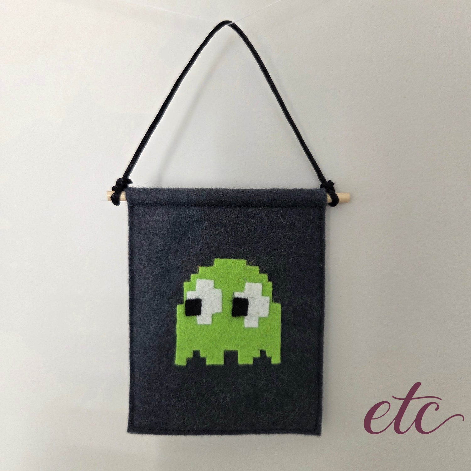 A dark charcoal gray felt wall hanging with a black hanging cord and a light wooden dowel, featuring a pixelated PacMan green ghost design on the front.