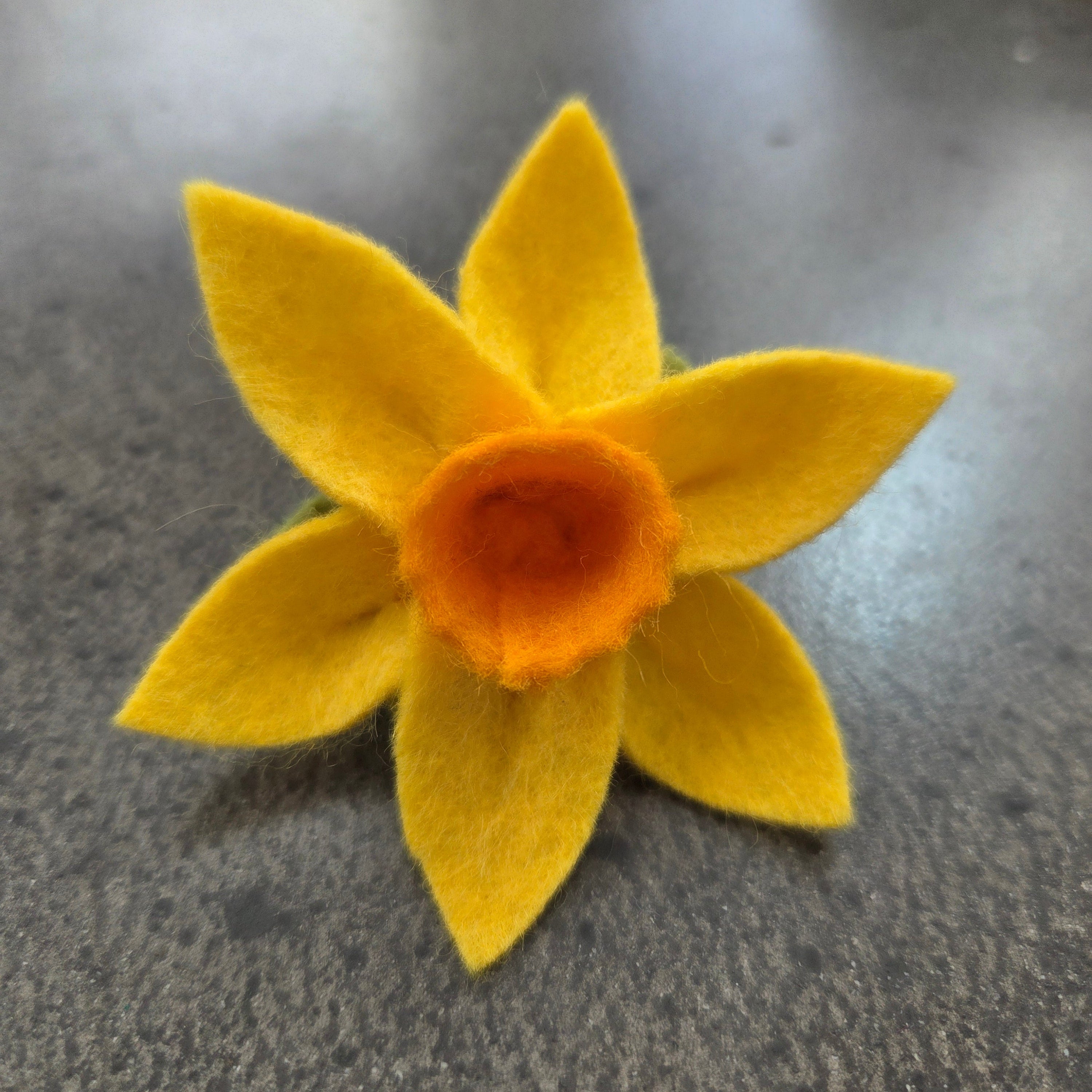 A handmade felt daffodil flower in bright yellow with a textured orange center, resting on a dark gray surface.