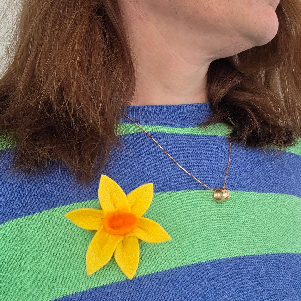 A close-up side profile of a woman with brown hair wearing a blue and green striped knit sweater, a gold necklace, and a yellow felt daffodil brooch on her chest.