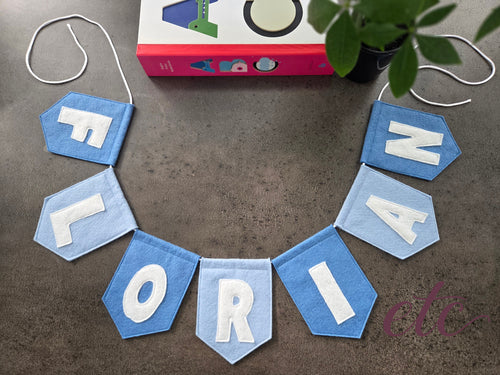 Felt name garland / bunting spelling 'FLORIAN' on a textured surface with a book and plant in the background.