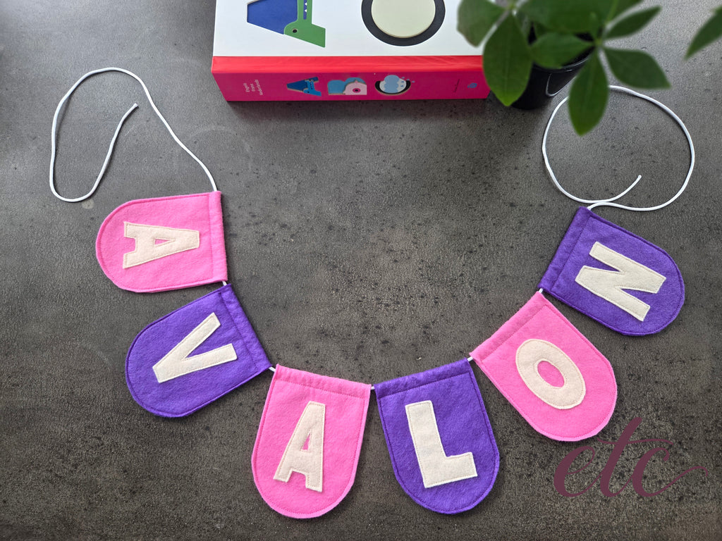 Felt letter garland / bunting spelling 'AVALON' on a dark surface with a book and plant in the background.