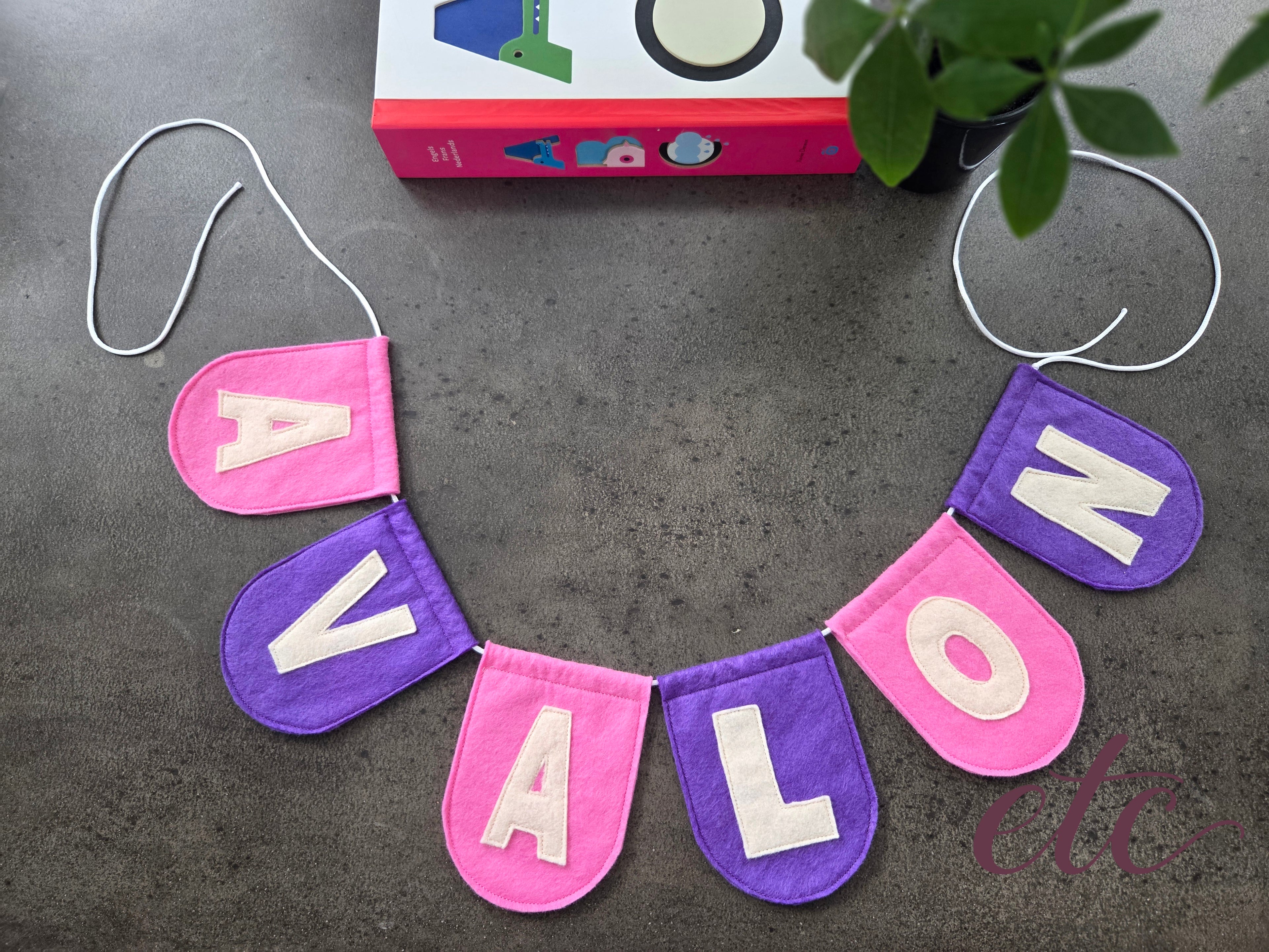 Felt letter garland / bunting spelling 'AVALON' on a dark surface with a book and plant in the background.