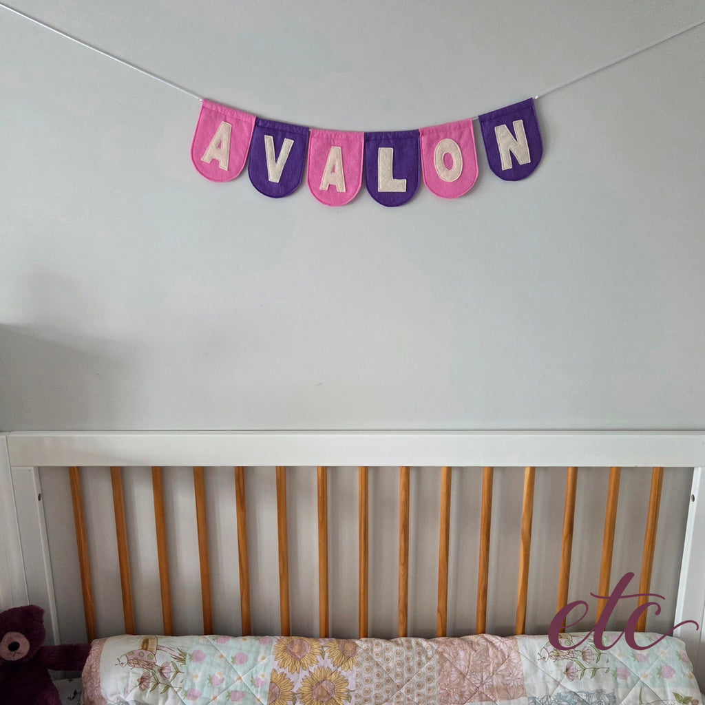 Personalised felt bunting / garland with 'Avalon' letters on a plain background above a crib.