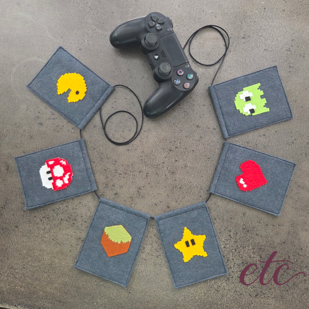A felt banner garland featuring pixel-art style icons from classic video games, including Pac-Man, a green monster, a Mario Bros red mushroom, a red heart, a yellow star, and a MineCraft green-and-brown box, strung on a black cord over a gray concrete background.