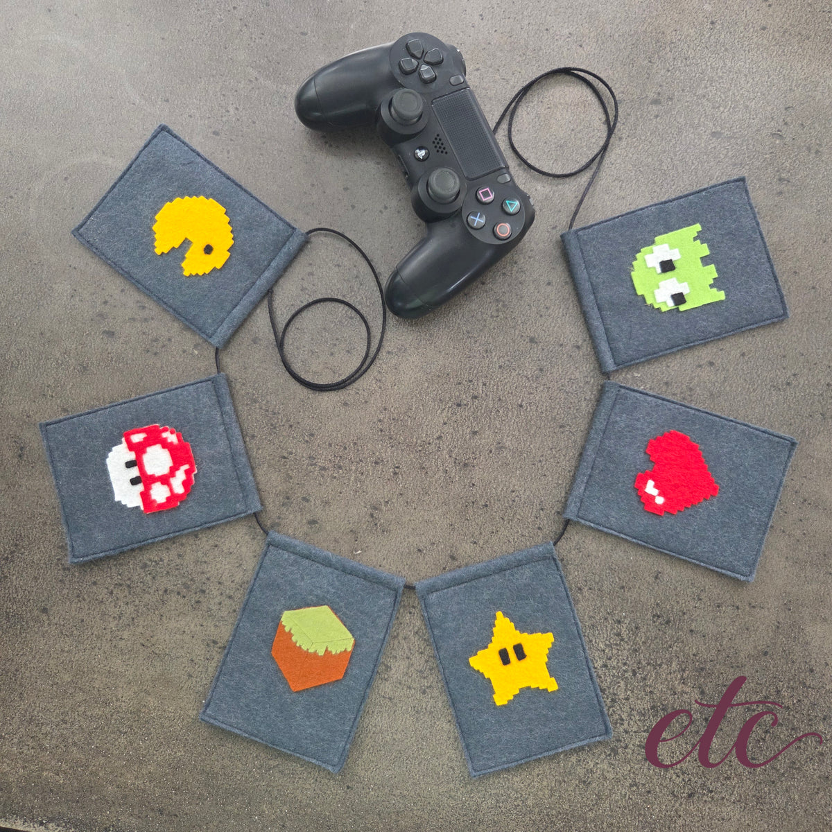 A felt banner garland featuring pixel-art style icons from classic video games, including Pac-Man, a green monster, a Mario Bros red mushroom, a red heart, a yellow star, and a MineCraft green-and-brown box, strung on a black cord over a gray concrete background.
