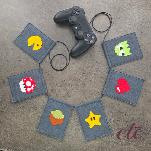 A felt banner garland featuring pixel-art style icons from classic video games, including Pac-Man, a green monster, a Mario Bros red mushroom, a red heart, a yellow star, and a MineCraft green-and-brown box, strung on a black cord over a gray concrete background.