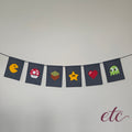 A string banner made of seven dark navy fabric flags, each featuring a different pixel-art video game icon including Pac-Man, a Mario Bros Mushroom, an MineCraft box, a Star, a Heart, and a Ghost. The banner is hung against a plain light gray wall.