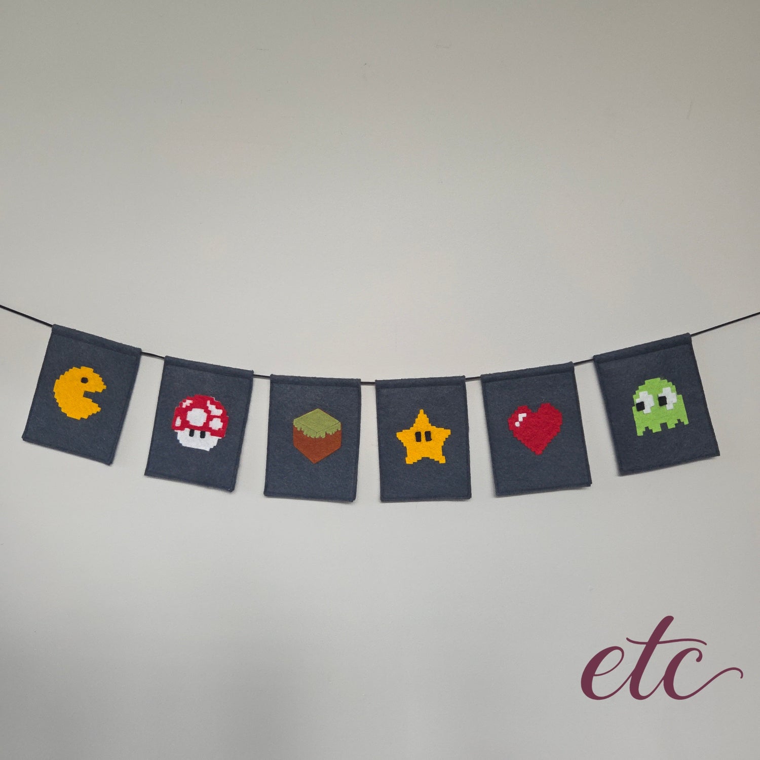 A string banner made of seven dark navy fabric flags, each featuring a different pixel-art video game icon including Pac-Man, a Mario Bros Mushroom, an MineCraft box, a Star, a Heart, and a Ghost. The banner is hung against a plain light gray wall.