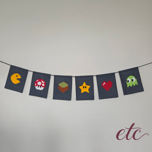 A string banner made of seven dark navy fabric flags, each featuring a different pixel-art video game icon including Pac-Man, a Mario Bros Mushroom, an MineCraft box, a Star, a Heart, and a Ghost. The banner is hung against a plain light gray wall.