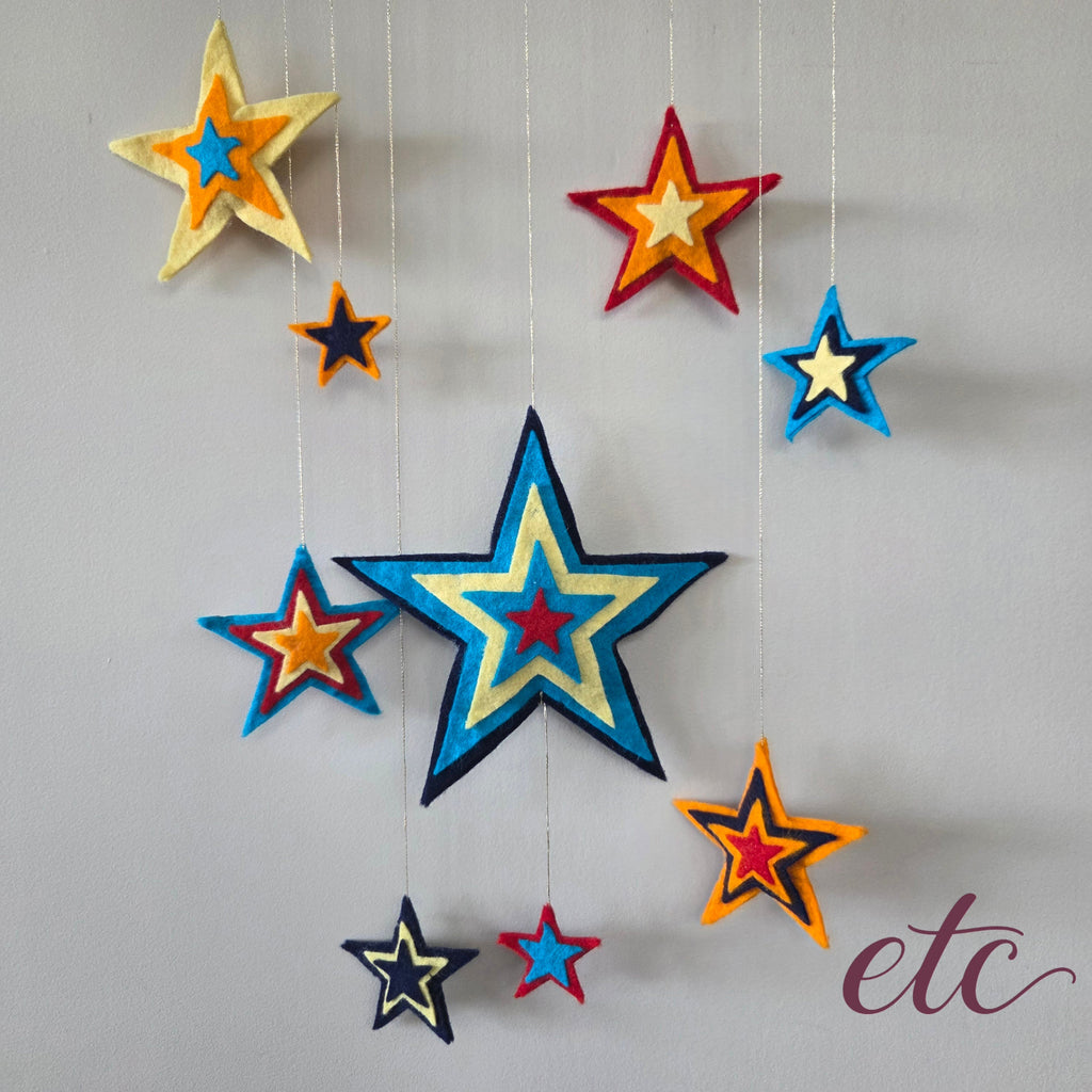 A set of colorful felt star ornaments in various sizes hanging from thin strings against a light gray wall. The stars feature layered colors including blue, yellow, red, orange, and navy, with one large blue star in the center and several smaller stars around it.